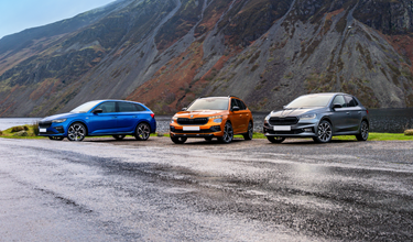 Four Škoda vehicles — including the Enyaq, Enyaq Coupé, and Kodiaq — displayed together on a forecourt outside a modern curved architectural building, surrounded by mature trees in a parkland setting at golden hour.